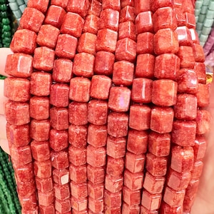 May include: Close-up of a strand of red-orange gemstone beads. The beads are cube-shaped with slightly rounded edges. The beads have a mottled appearance, with variations in color and texture. The beads are strung together, ready for jewelry making.