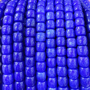 May include: Close-up of a strand of vibrant blue beads. The beads are a rounded square shape and have a glossy finish. The beads are strung together, creating a dense, textured pattern. The color is a deep, saturated blue.