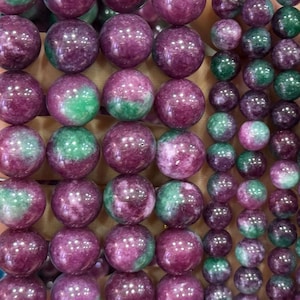 May include: Close-up of various gemstone beads. The beads are primarily round, with a mix of deep purple and green hues. Some beads are solid purple, while others have a green section. The beads are arranged in rows, suggesting they are for jewelry making.