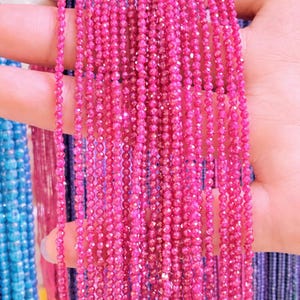 May include: Close-up of multiple strands of bright pink gemstone beads. The beads are small, faceted, and strung together, creating a shimmering effect. The strands are held in a hand, with other strands of blue beads visible in the background.