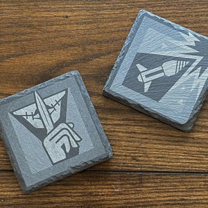 May include: Two square slate coasters with etched designs. One shows a hand holding a knife, the other a rocket and drill. The coasters are grey with white etched graphics, set on a wooden surface. These coasters are ideal for home decor.