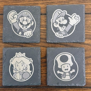 Könnte beinhalten: Set aus vier quadratischen Schieferuntersetzern mit Charakteren aus der Super Mario-Videospielserie. Jeder Untersetzer zeigt einen anderen Charakter: Mario, Luigi, Prinzessin Peach und Toad, alle in einem weißen und grauen Design.