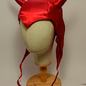 Könnte beinhalten: Ein roter Satin-Teufelshut mit spitzer Front und langen, fließenden Bändern. Der Hut wird auf einem Schaufensterkopf präsentiert, um seine Form und sein Design zu zeigen. Die Hörner sind gebogen und stehen aufrecht.