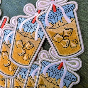 May include: Embroidered patches featuring iced coffee drinks. Each patch has a blue lid, a red straw, and a light pink bow. The drink is a light brown color with ice cubes. The patches are on a green surface.