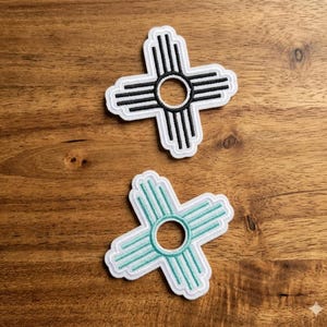 May include: Two embroidered patches featuring the Zia symbol, a Native American design. One patch has a black design with a white border, and the other has a teal design with a white border. The patches are on a wooden surface.