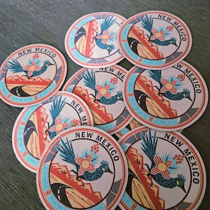 May include: A collection of round stickers featuring a roadrunner bird, a symbol of New Mexico. The stickers have a beige background with a turquoise border and the words "NEW MEXICO". The design includes a road, mountains, and a sun symbol.