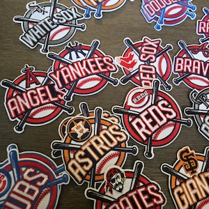 May include: A collection of embroidered baseball team patches featuring various logos, team names, and crossed bats over a baseball graphic. The patches include the Mets, Dodgers, White Sox, Yankees, Angels, Red Sox, Braves, Astros, Reds, Cubs, Pirates, and Giants.