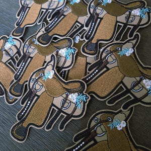 May include: Embroidered patches featuring a horse saddle design. The patches have a tan and brown color scheme, with accents of white and light blue floral details. The patches are likely for clothing or accessories.