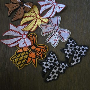 May include: A collection of embroidered bow-shaped patches featuring various sports themes, including a brown football bow, a yellow softball bow, white baseball bows with red stitching, an orange basketball bow, and black and white soccer ball bows.