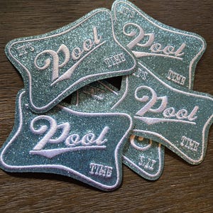 May include: A collection of glittery teal patches with the words "It's Pool Time" embroidered in white script. The patches have a unique, curved shape and are scattered on a wooden surface.