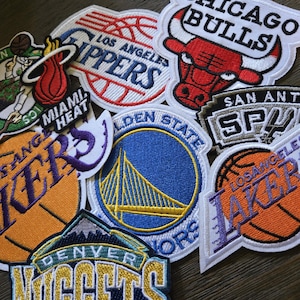 May include: A collection of embroidered patches featuring logos of various NBA basketball teams, including the Chicago Bulls, Los Angeles Lakers, and Golden State Warriors. The patches are in various colors and shapes, with detailed stitching.