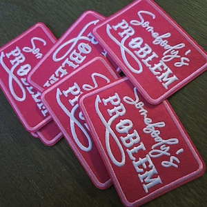 May include: Several pink rectangular patches with white embroidered text that reads "Somebody's PROBLEM" in a cursive font. The patches are arranged in a slightly overlapping pattern, with a pink border.