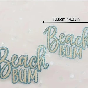 May include: Two glittery blue and white "Beach Bum" cutouts. The cutouts are approximately 4.25 inches wide and 2.87 inches tall. The text is in a cursive font.