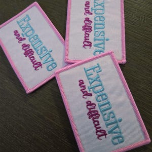 May include: Three rectangular patches with the embroidered text "Expensive and difficult". The patches have a light pink border and the text is in blue and pink. The patches are on a dark brown surface.