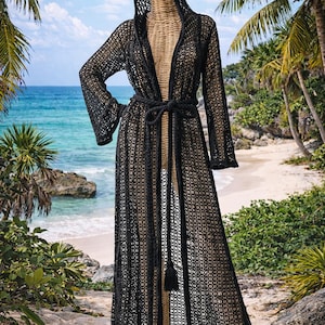 May include: A black, open-weave hooded robe with long sleeves and a matching belt. The robe is floor-length and features a tassel at the waist. The background shows a beach scene with palm trees and the ocean.