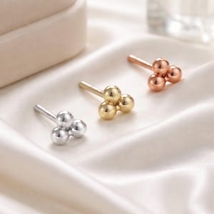 May include: Three stud earrings in silver, gold, and rose gold. Each earring features three small, spherical beads arranged in a triangular cluster. The earrings are displayed on a white, silky fabric.