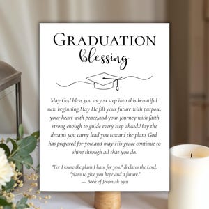 May include: A white graduation blessing sign with the words "GRADUATION blessing" in elegant script. The sign includes a graduation cap illustration and a religious quote about hope and the future. A candle and floral arrangement are in the background.
