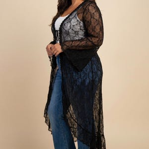 May include: A long, black lace duster cardigan featuring long, flared sleeves and a delicate floral pattern throughout the fabric. The garment has an open front design with a thin tie closure at the neckline and a scalloped hemline finish.