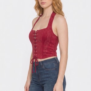 May include: A deep red, sleeveless, cropped halter top featuring a front lace-up closure with matching red cord ties. The garment has a square neckline and subtle, tonal embroidered flame-like patterns on the side panels, paired with dark blue denim jeans.