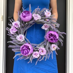 May include: A circular wreath adorned with large, deep purple peonies and delicate, pale purple blooms. The wreath's foundation is made of grey-green foliage, providing a beautiful contrast. This floral wreath is ideal for adding a touch of elegance to any space.