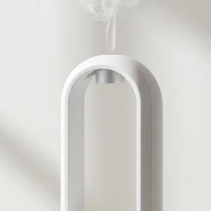 May include: White aromatherapy diffuser with a digital display showing "99". The device has a rounded base and a tall, arched top. Wisps of vapour rise from the top. A sprig of green leaves is in the lower left.