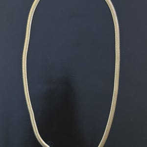 Könnte beinhalten: Eine goldfarbene Halskette mit Fischgrätenmuster. Die Kette besteht aus einer Reihe kleiner, ineinandergreifender Glieder, die ein strukturiertes, schlangenartiges Aussehen erzeugen. Der Verschluss ist ebenfalls goldfarben. Die Kette wird vor einem dunklen Hintergrund präsentiert.