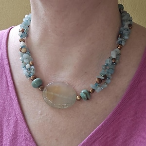 Könnte beinhalten: Eine mehrsträngige Halskette mit einem großen, ovalen, durchscheinenden beigen und hellgrünen Steinanhänger. Die Kette besteht aus kleinen, getrommelten hellblauen Steinchips, metallisch bronzefarbenen Perlen und winzigen dunkelblauen Glasakzenten.