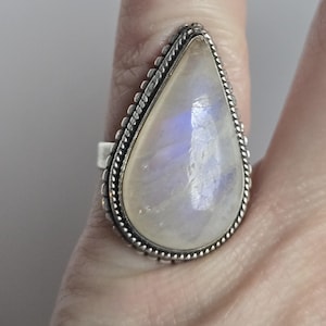 Könnte beinhalten: Ein großer, tropfenförmiger Mondsteinring. Der Edelstein ist ein blasses, durchscheinendes Weiß mit dezentem blauem und violettem Schimmer. Der Ring hat einen silberfarbenen, perlenbesetzten Rand und ein schlichtes Band.