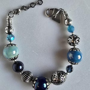 Könnte beinhalten: Ein Armband mit verschiedenen blauen Perlen, darunter runde, facettierte und dekorative Silberperlen. Es hat einen Silberverschluss und einen kleinen Anhänger. Die Perlen reichen von hellblau bis dunkelmarineblau. Ca. 19 cm lang.