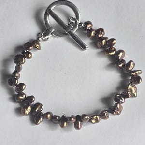 Könnte beinhalten: Ein Armband mit unregelmäßig geformten, bronzefarbenen Perlen. Die Perlen sind aufgereiht und mit einem silberfarbenen Knebelverschluss befestigt. Das Armband wird vor einem weißen Hintergrund gezeigt. Die Perlen haben einen Glanz.