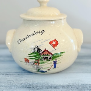 Puede incluir: Tarro de cerámica color crema con tapa, con una ilustración escénica de un pueblo suizo con una bandera roja y blanca. La palabra "Beatenberg" está escrita en el tarro.