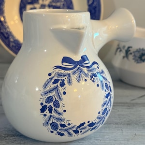 Puede incluir: Cremera de cerámica blanca con un diseño de corona navideña azul. La cremera tiene un pico y un asa. Un lazo azul está en la parte superior de la corona. Un plato azul y blanco está en el fondo.