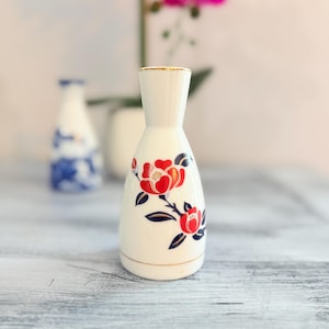 Puede incluir: Una botella de sake de cerámica blanca con un diseño floral rojo y ribete dorado. La botella tiene un cuello delgado y un cuerpo redondeado. Una botella similar está en el fondo, junto con una flor rosa.
