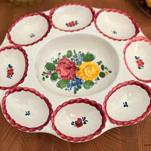 May include: White ceramic serving platter with eight oval compartments and a central bowl. The rim of each compartment is trimmed with a red rope design. The centre bowl features a floral design with red, yellow, and blue flowers.