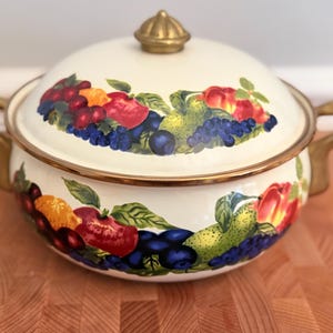 May include: Cream-colored enamelware pot with a lid, adorned with a vibrant fruit design featuring apples, grapes, and pears. The pot has gold-colored handles and a gold knob on the lid. A decorative kitchenware item.