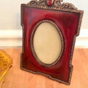 Puede incluir: Un marco de fotos decorativo rojo con una abertura ovalada. El marco tiene detalles dorados ornamentados alrededor de los bordes y la parte superior. El marco está sobre una superficie de madera.