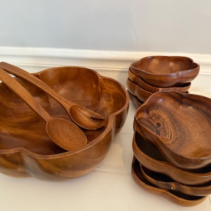 Pode incluir: Um conjunto de saladeiras de madeira com uma tigela grande para servir, dois utensílios de servir de madeira e várias tigelas individuais menores. As tigelas e os utensílios são feitos de madeira de tom médio e quente.