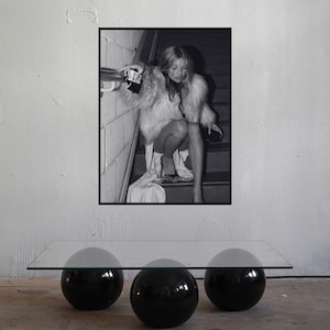 May include: A rectangular glass coffee table featuring three large, glossy black spherical bases. Above the table, a framed black and white photograph depicts a person in a fur coat sitting on a staircase, holding a small clutch and wearing heels.