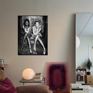 Könnte beinhalten: Schwarz-Weiß-Fotografie von zwei rauchenden Frauen, gerahmt. Das Bild hängt an einer beigefarbenen Wand in einem Wohnzimmer. Eine runde Lampe, Bücher und eine Kerze sind ebenfalls zu sehen.