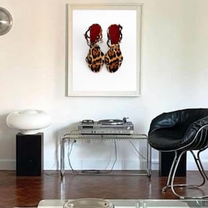 May include: A pair of open-toe sandals featuring a brown and black leopard print pattern, thin ankle straps with buckles, and a solid red insole, displayed within a white rectangular frame hanging on a plain wall above a metal console table.