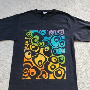 May include: Black t-shirt featuring a vibrant, abstract design. The design is a square of colorful, organic shapes in shades of yellow, orange, green, blue, and purple. The shirt is made of a soft material and has short sleeves.