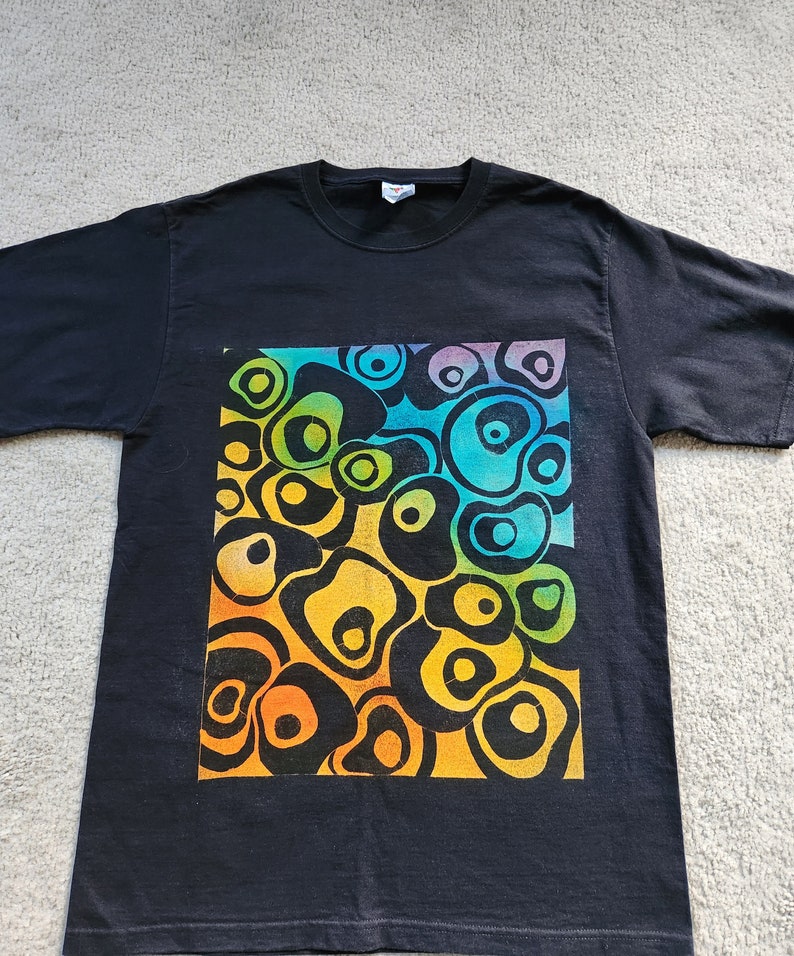 May include: Black t-shirt featuring a vibrant, abstract design. The design is a square of colorful, organic shapes in shades of yellow, orange, green, blue, and purple. The shirt is made of a soft material and has short sleeves.