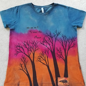 May include: A short-sleeved t-shirt with a colorful design. The top portion is blue, transitioning to pink and orange at the bottom. Black tree silhouettes are featured, with the text "We ran as if To meet the Moon." A small patch with a raven is at the bottom.