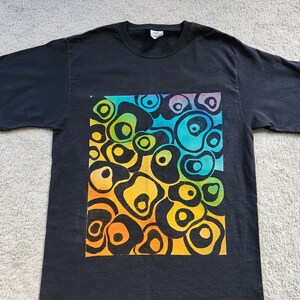 May include: Black t-shirt featuring a vibrant, colorful design of abstract, organic shapes. The design transitions through shades of yellow, orange, green, and blue, creating a visually striking pattern. The shirt is a classic crew neck style.