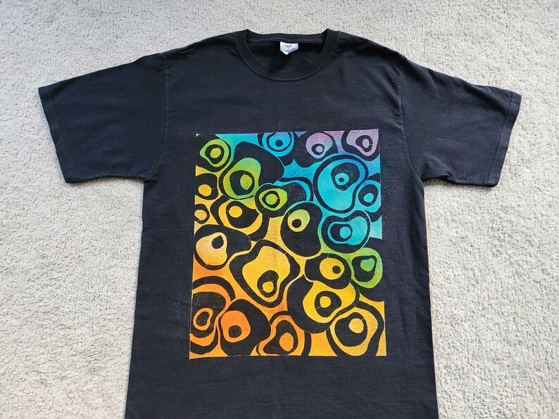 May include: Black t-shirt featuring a vibrant, colorful design of abstract, organic shapes. The design transitions through shades of yellow, orange, green, and blue, creating a visually striking pattern. The shirt is a classic crew neck style.