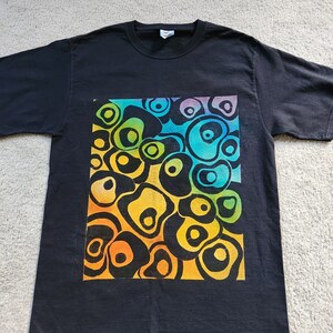 May include: Black t-shirt featuring a vibrant, abstract design. The design is a colorful arrangement of interconnected, organic shapes in shades of orange, yellow, green, blue, and purple. The t-shirt is made of a soft material and has a classic crew neck.