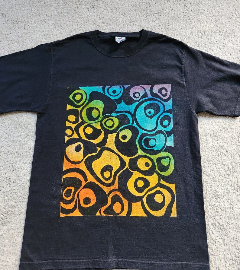 May include: Black t-shirt featuring a vibrant, abstract design. The design is a colorful arrangement of interconnected, organic shapes in shades of orange, yellow, green, blue, and purple. The t-shirt is made of a soft material and has a classic crew neck.
