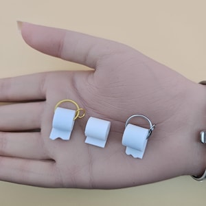 May include: Three miniature white toilet paper roll charms. One charm has a gold-colored ring, and the other has a silver-colored ring. The third charm is a small white toilet paper roll without a ring. The charms are displayed on a light skin-toned background.