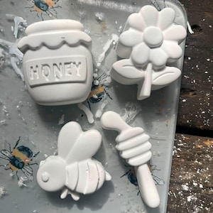 Puede incluir: Cuatro figuras blancas de yeso o cera que incluyen un tarro de miel con la palabra HONEY en relieve en el frente, una flor con pétalos redondeados, una abeja con alas y un cazo para miel. Los objetos están dispuestos sobre una superficie ilustrada.