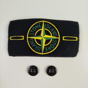 Może przedstawiać: Prostokątna czarna naszywka z wyhaftowanym żółto-zielonym logo kompasu z napisem Stone Island. Naszywka posiada dwa żółte poziome paski i jest uzupełniona dwoma małymi czarnymi okrągłymi guzikami z czterema otworami.
